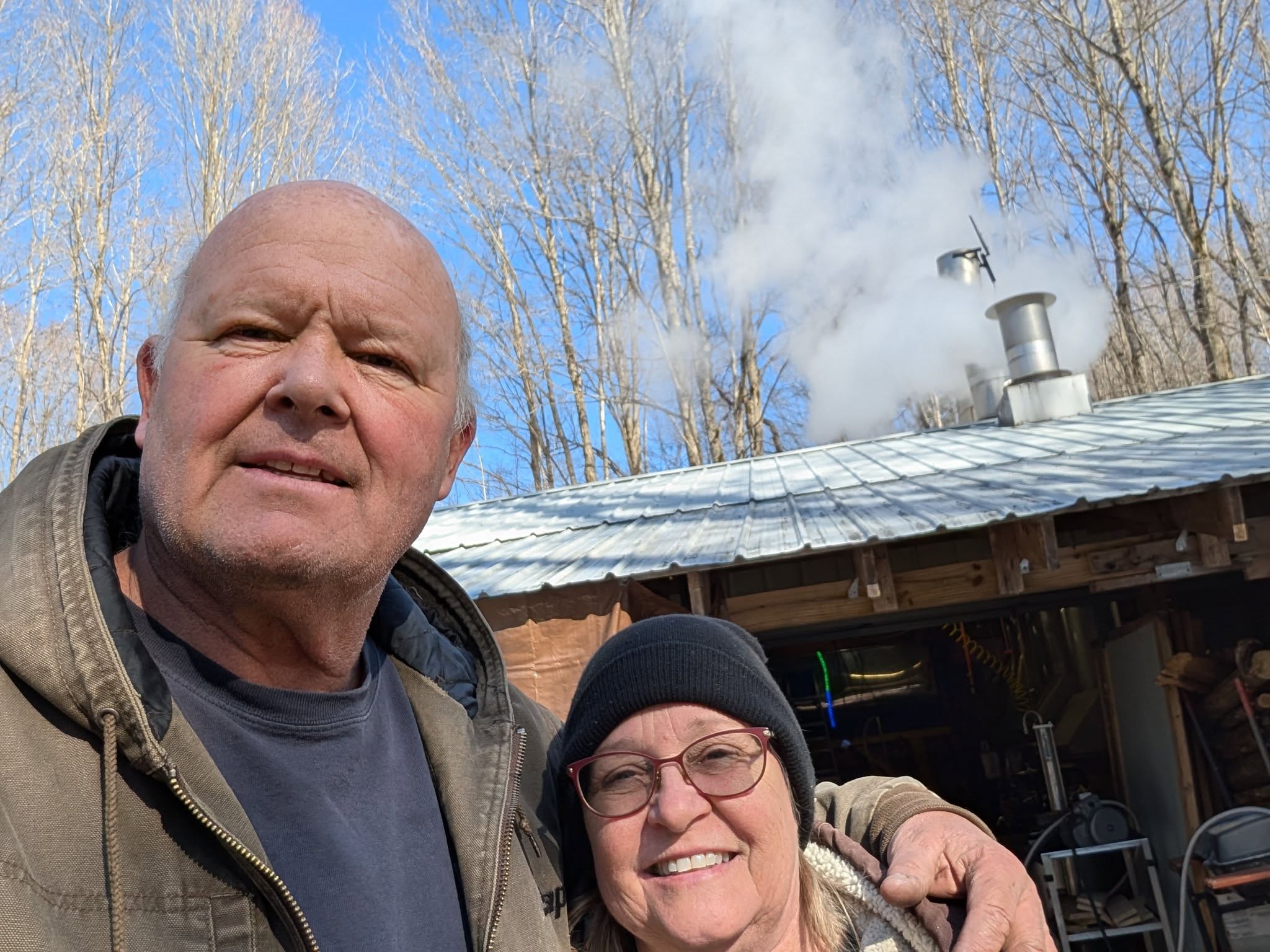 Ron and Kenna Rhynard outside the sugar camp, steam rising from the evaporator stack behind them